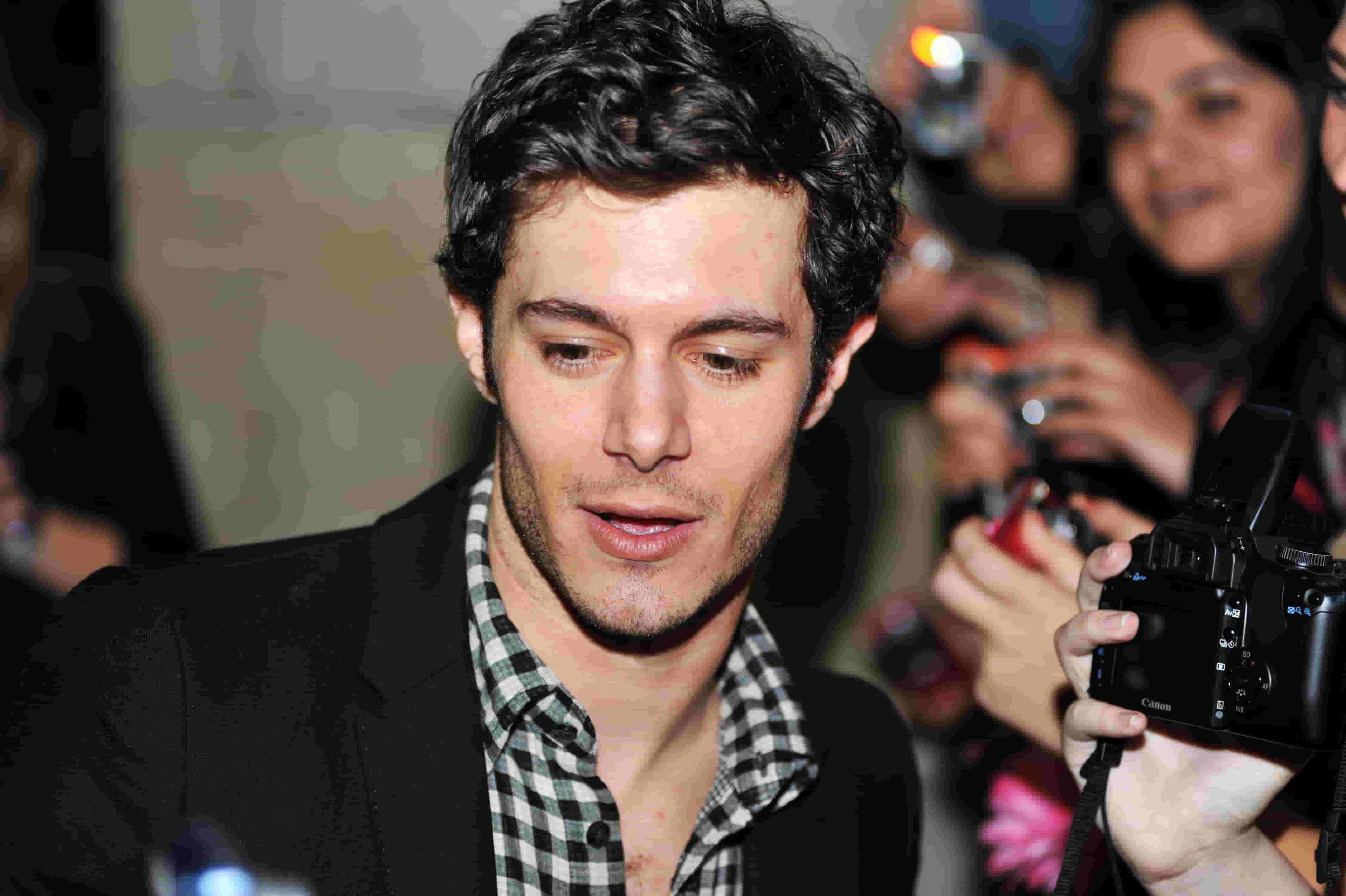 Adam Brody wearing a black and white shirt under a black coat at an event, surrounded by photographers.