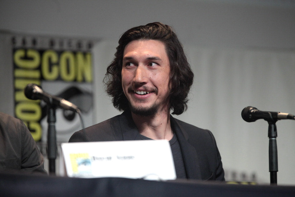 Adam Driver in a black suit, looking towards the side and smiling.