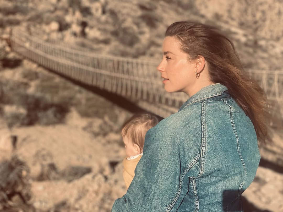 Amber Heard, wearing a blue denim shirt, holds her child as the wind blows through her hair.