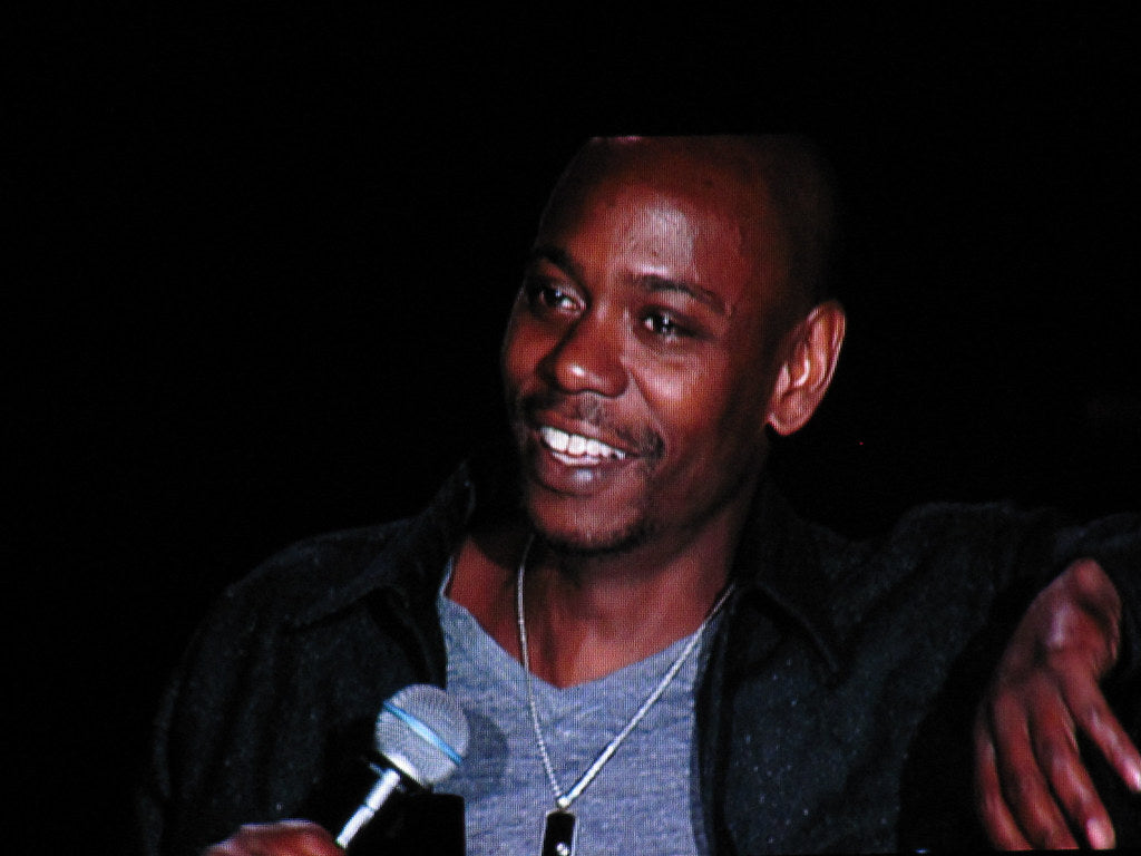 A close-up of Dave Chappelle smiling, wearing a gray shirt with a black jacket over it and a silver chain with a pendant around his neck, while holding a microphone in his right hand.