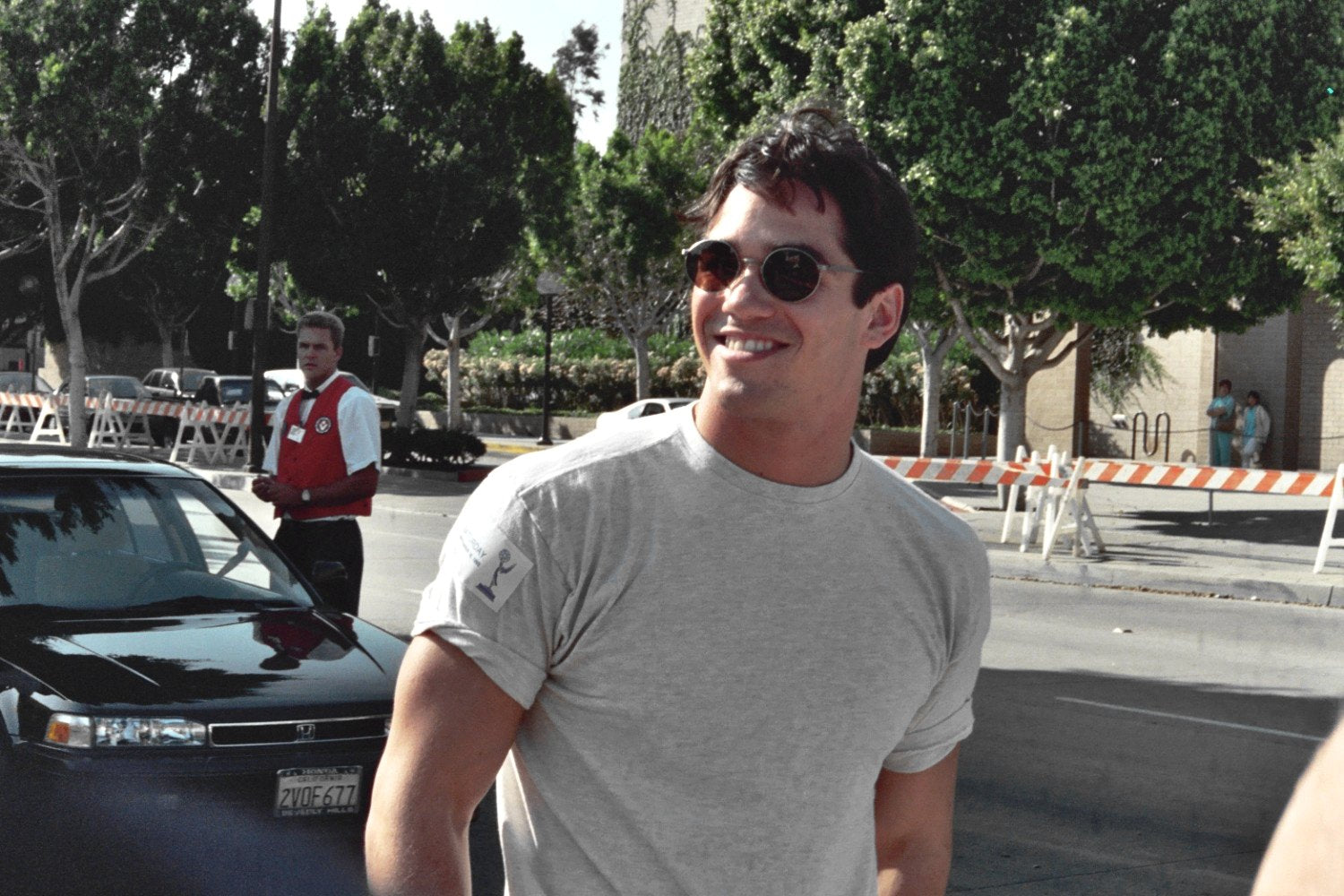 Dean Cain in a grey t-shirt and black shades, smiling casually while walking on the road.