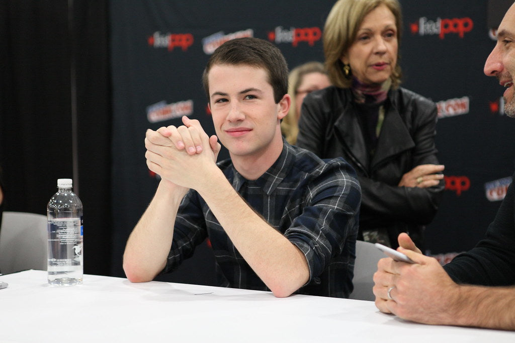 Dylan Minnette wearing a black checkered shirt, with his hands clasped and smiling at the camera