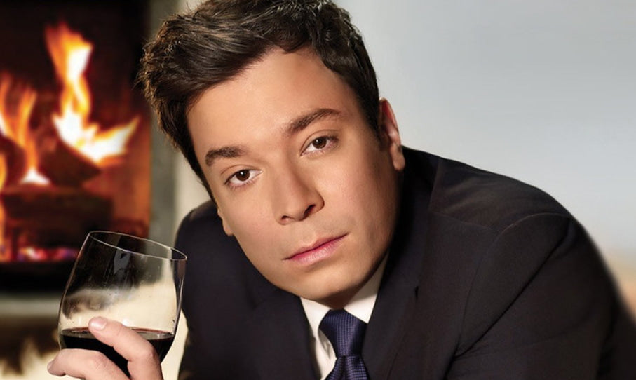 Front close-up of Jimmy Fallon, in a black suit, holding a glass of red wine in his hand.
