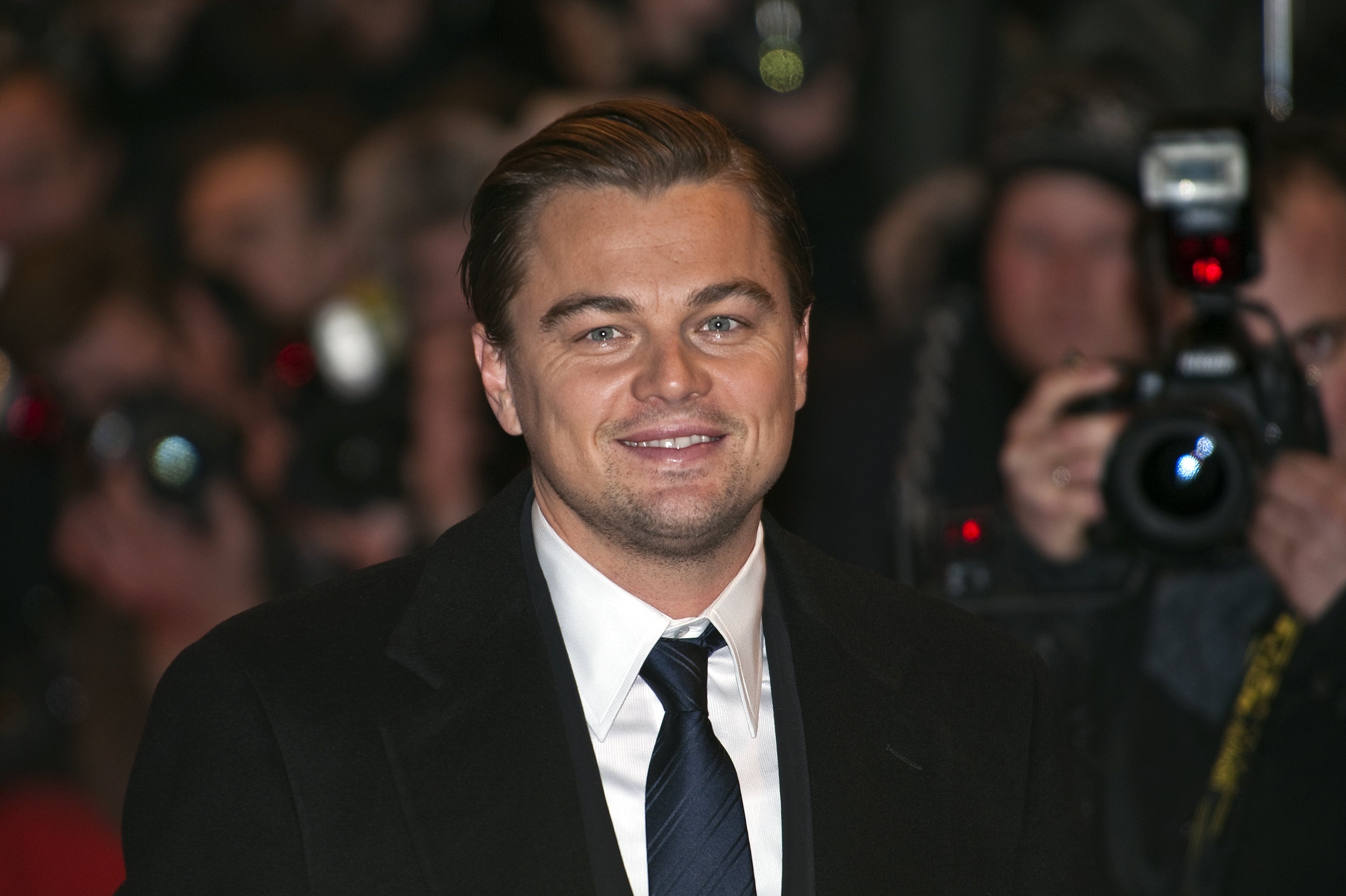 Leonardo DiCaprio looking composed, dressed in a sharp black suit, standing in front of paparazzies.