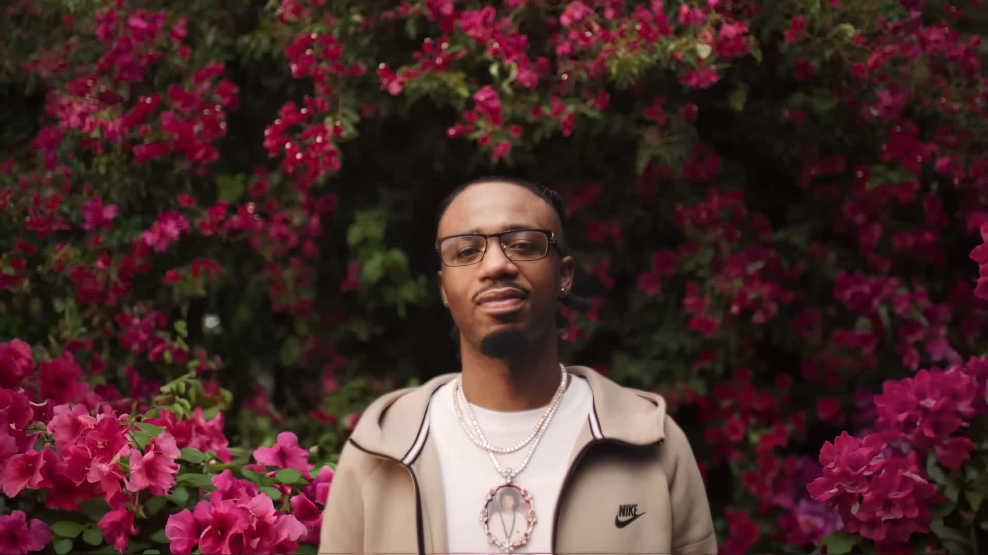 Metro Boomin wearing a Nike light brown jacket, standing in front of pink bougainvillea trees