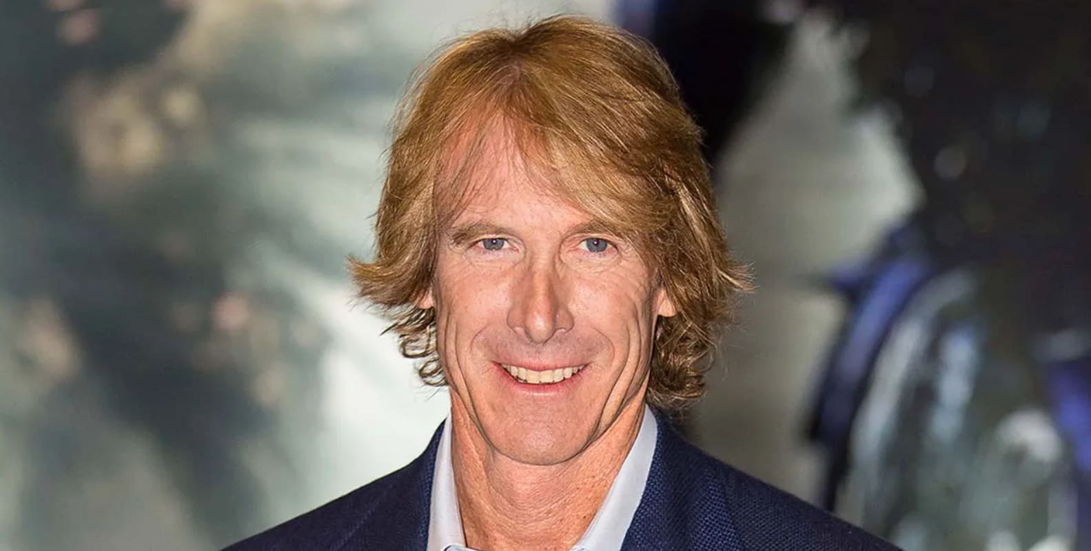 Michael Bay in a blue suite and white shirt, smiling while looking at the camera.