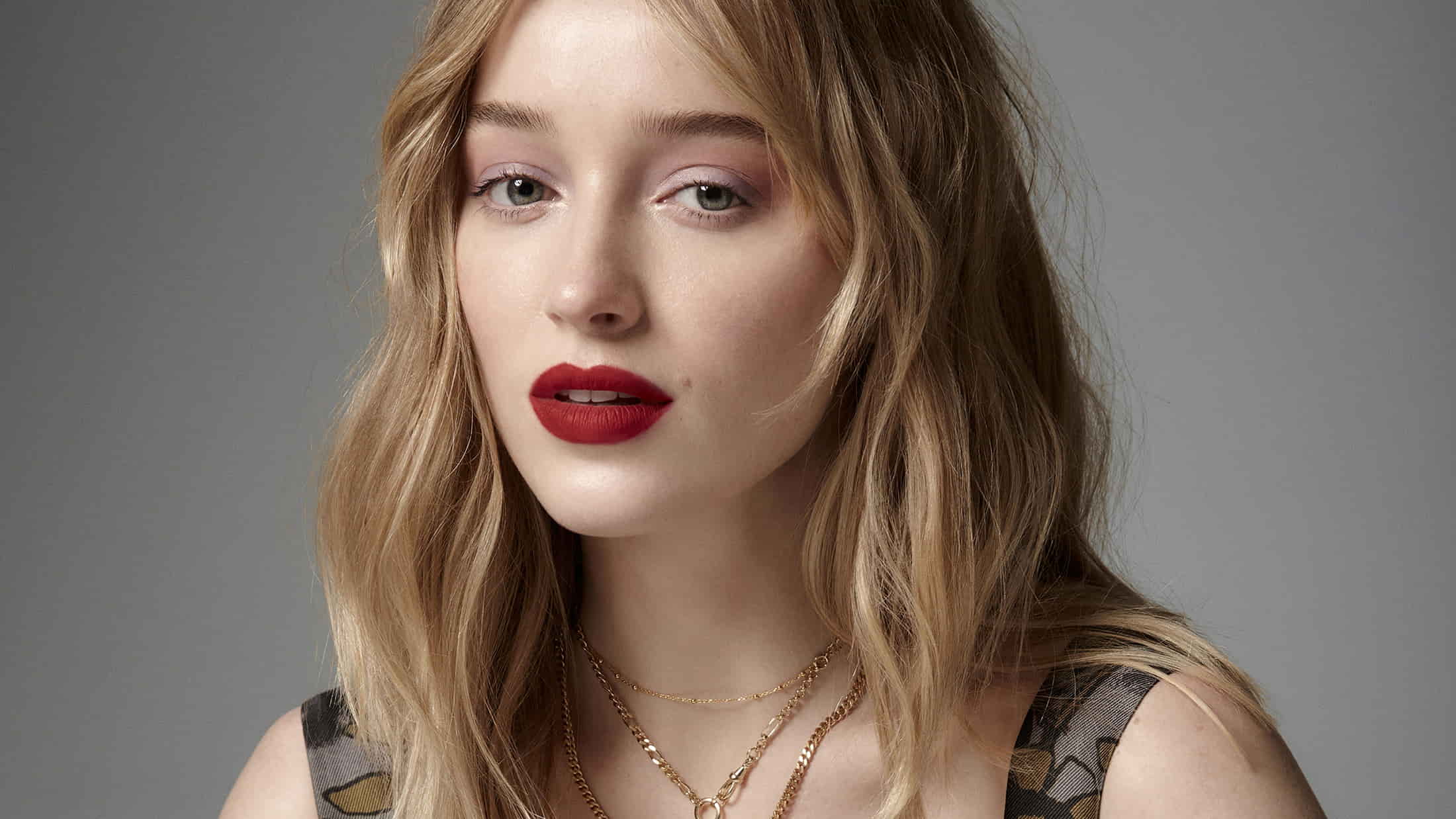 Phoebe Dynevor with wavy blonde hair, bold red lips, and layered gold necklaces against a neutral backdrop.