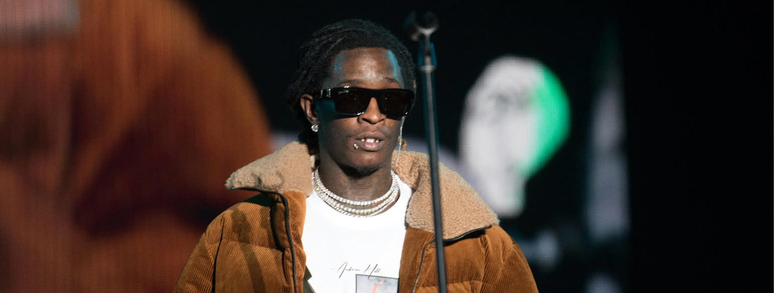 Young Thug, wearing black shades and a brown puffer jacket over a white t-shirt at his concert.