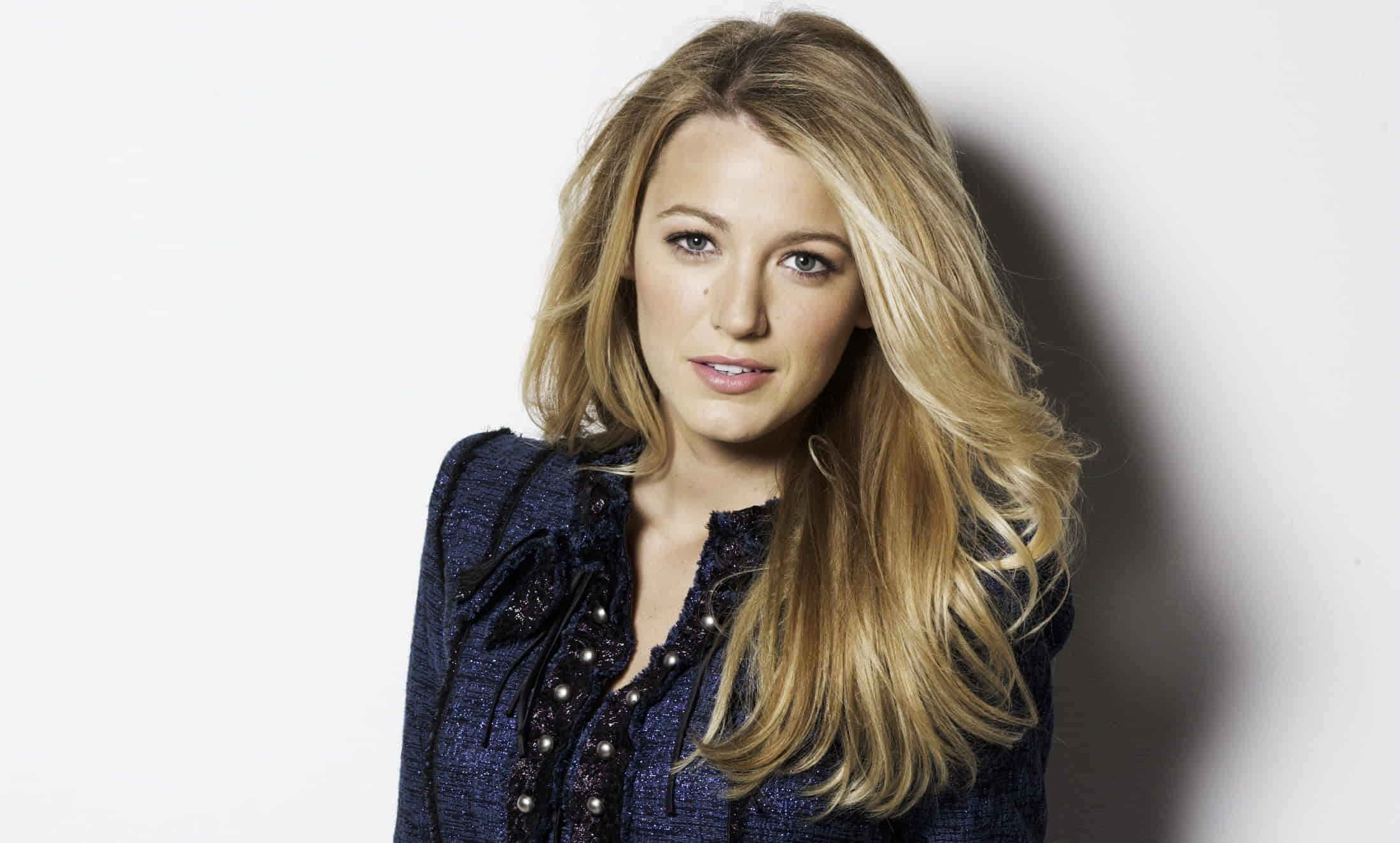 A portrait of Blake Lively with long, wavy blonde hair, wearing a navy blue textured jacket with pearl embellishments, against a neutral background.