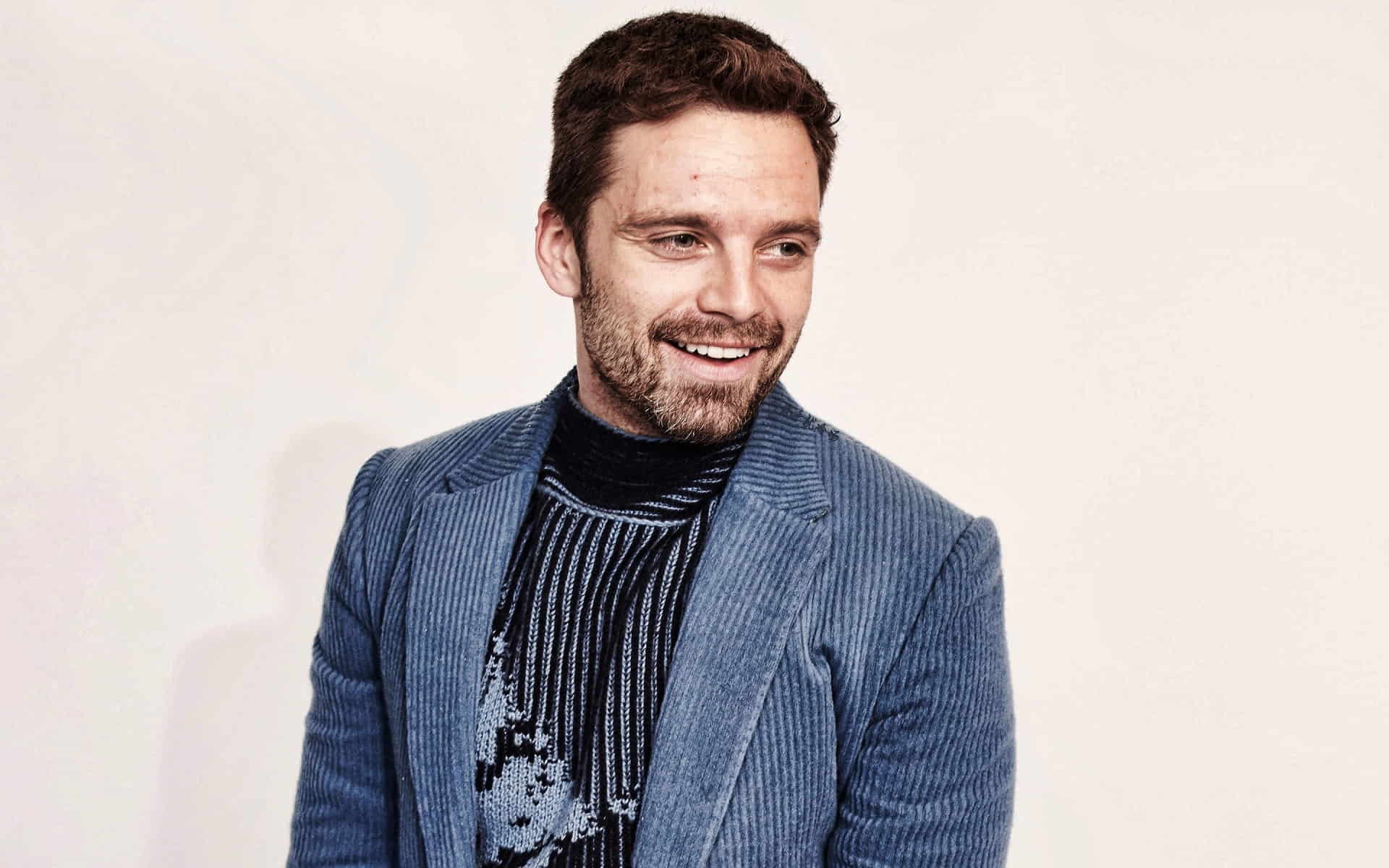 Sebastian Stan in a blue jacket and turtleneck sweater smiling while looking to the side.