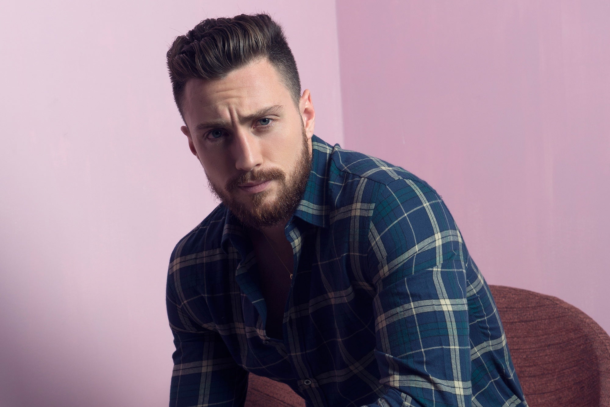 Aaron Tylor in an indigo-colored check shirt, posing for the camera, in front of a pink background