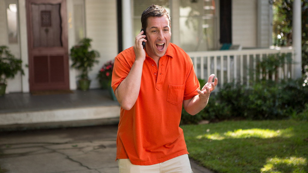 Adam Sandler in a bright orange t-shirt, looking amazed while talking to someone over call