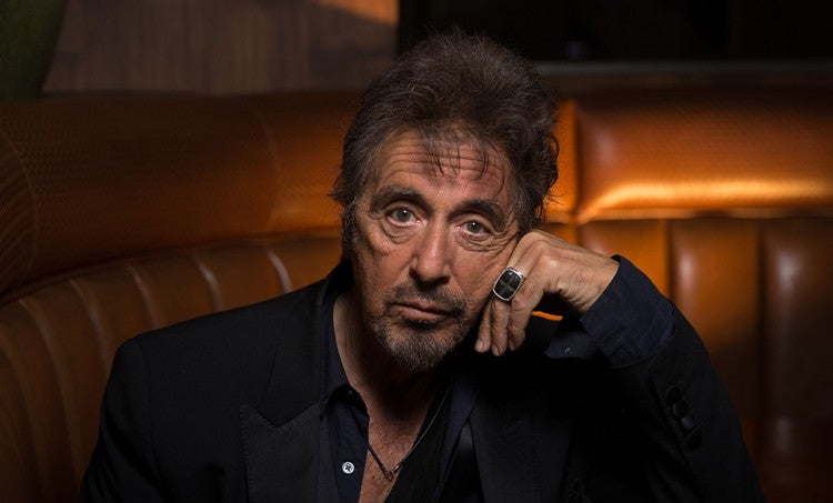 Al Pacino in a black suit, sitting in front of an orange couch, posing with his left hand on his cheek.