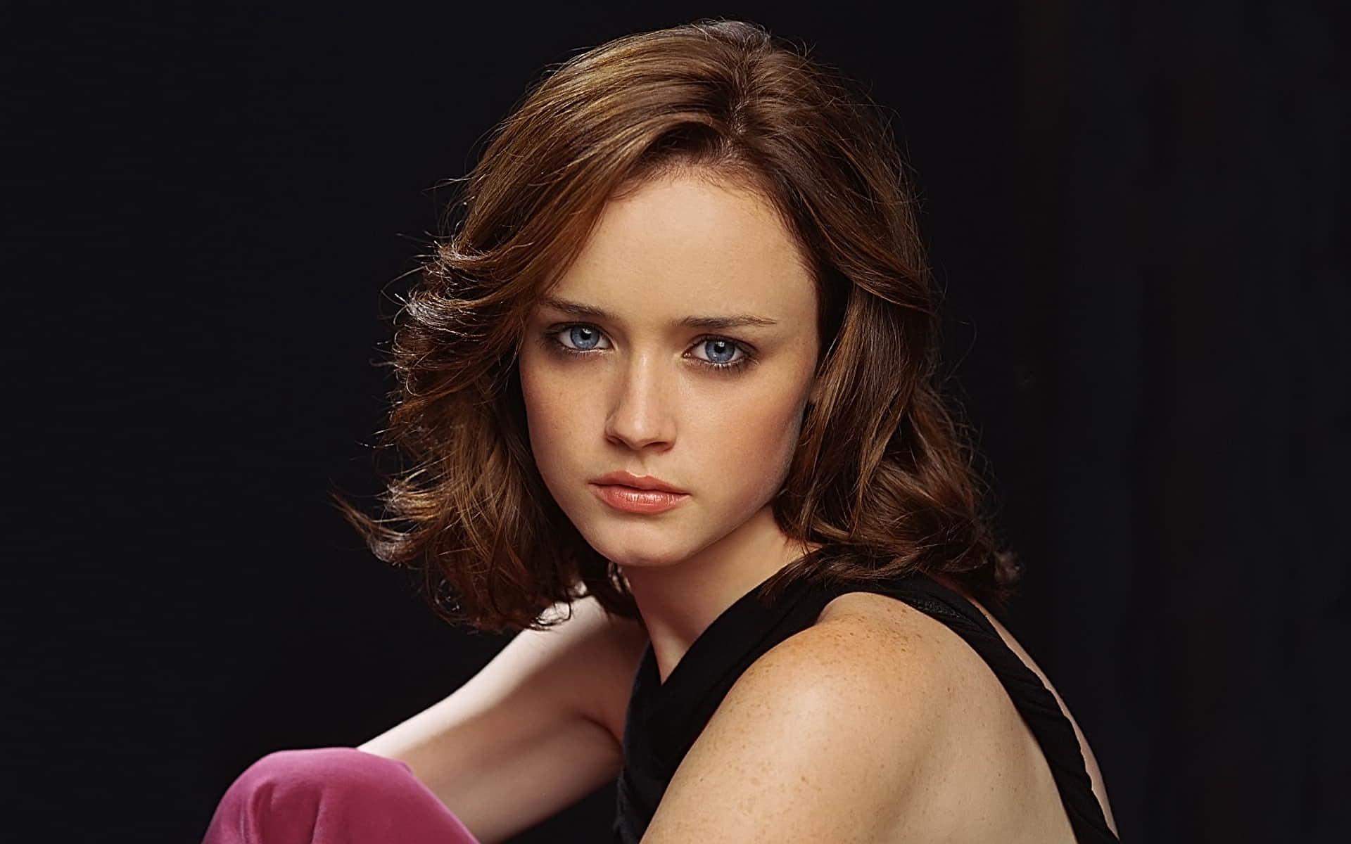 Alexis Bledel in a black dress and open hair, posing in front of a black background.