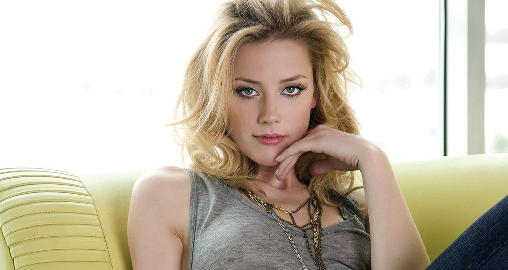 Amber Heard is sitting on a yellow sofa wearing a grey top, holding her hand near her cheek and looking at the camera