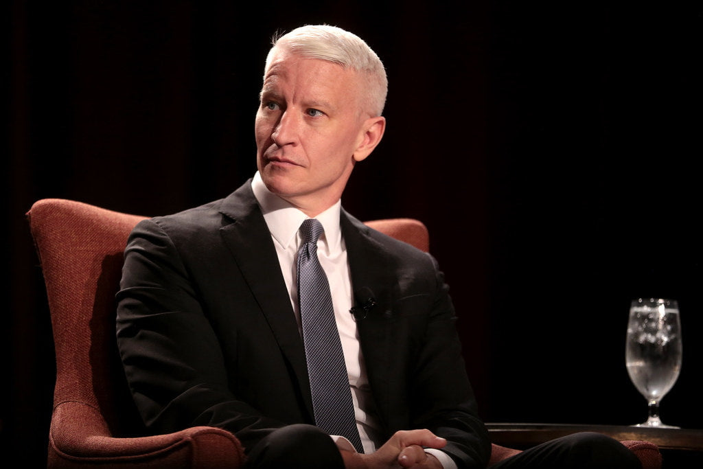 Anderson Cooper, in a black suit, white shirt, and gray tie, is sitting on a red couch at a conference