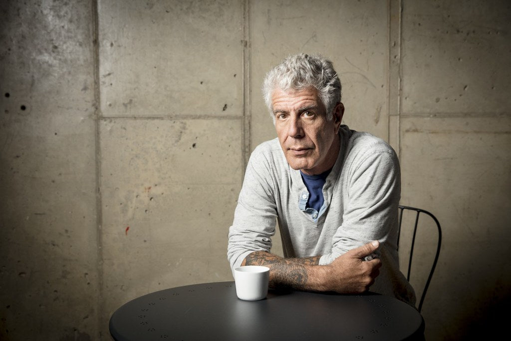 Anthony Bourdain in a grey t-shirt, sitting on a chair and posing for the camera in front of a cream brick wall