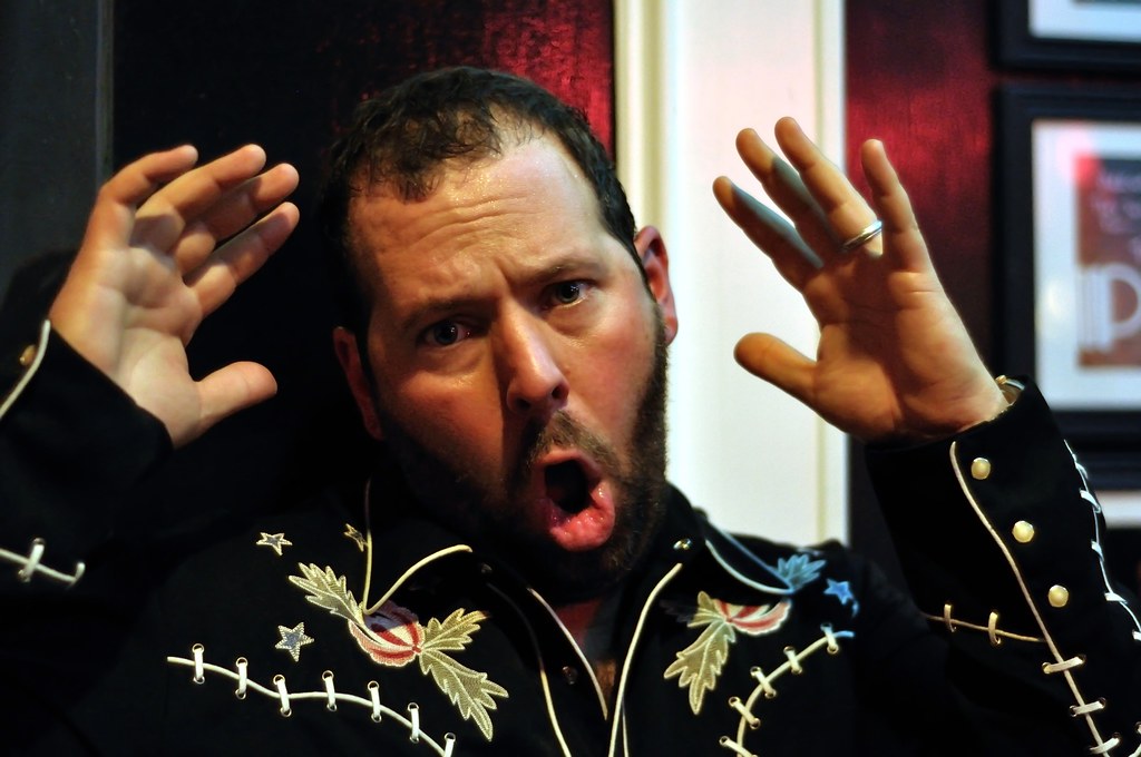 Bert Kreischer has his mouth open and hands raised. He appears to be making a gesture or sign with his hands.