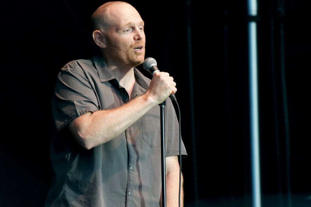 Bill Burr in a grey shirt, holding a microphone in his right hand while addressing the crowd