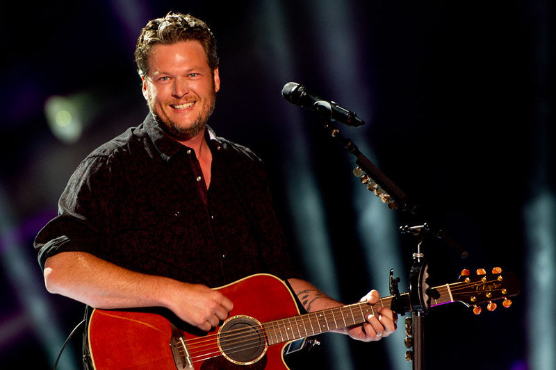 Country music singer Blake Shelton is smiling and playing guitar on stage with a red and black button up shirt and dark jeans.