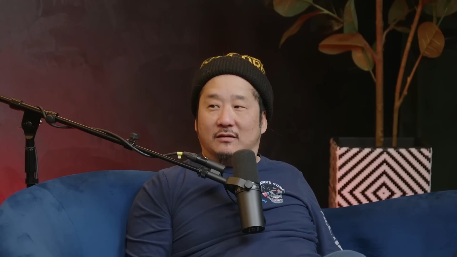 Bobby Lee at a podcast, wearing a blue t-shirt and a beanie cap, sitting with a microphone kept in front of him.