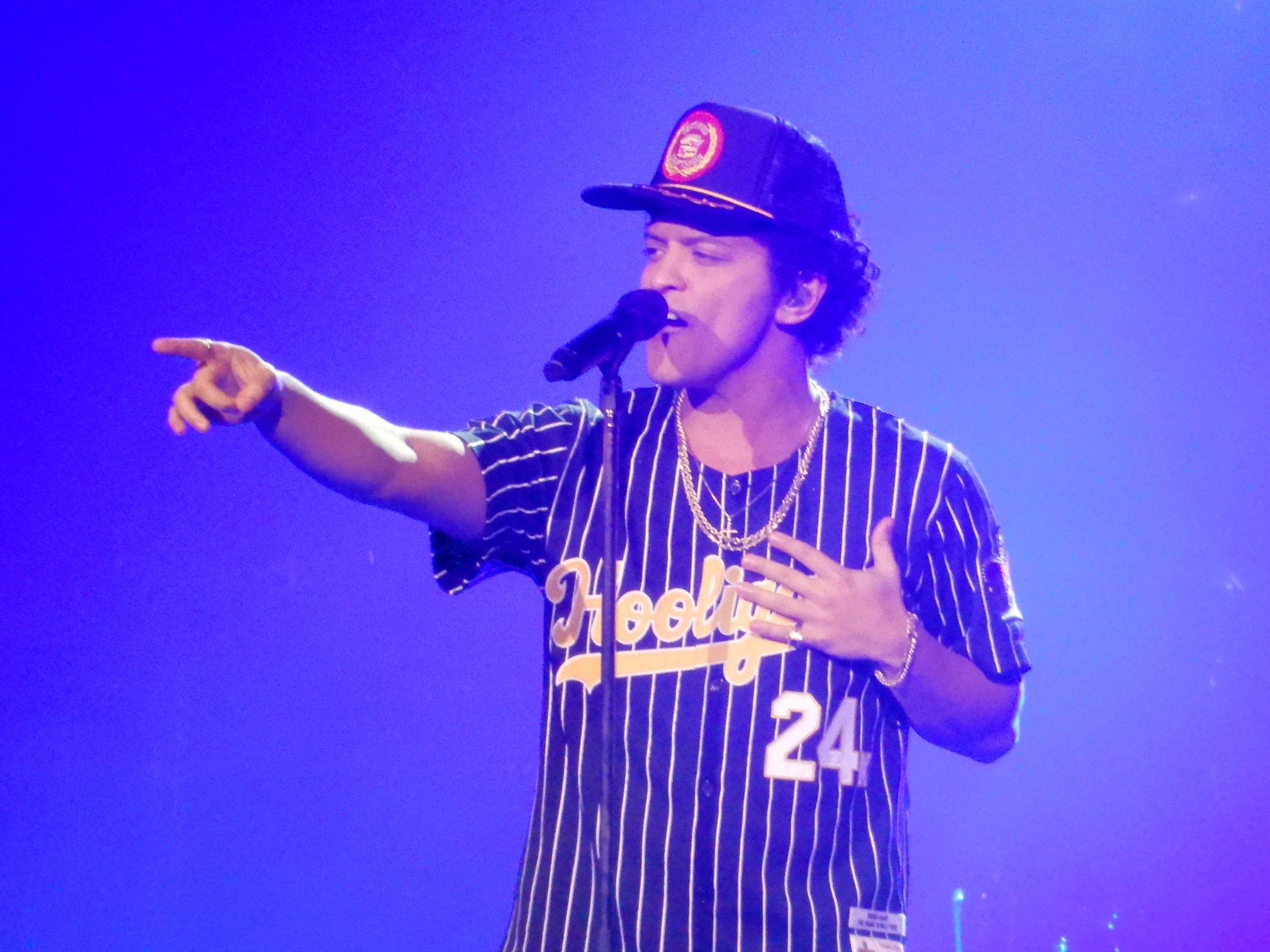 Bruno Mars is on the stage, performing with a microphone in front of him. He is pointing his hand towards the right side.