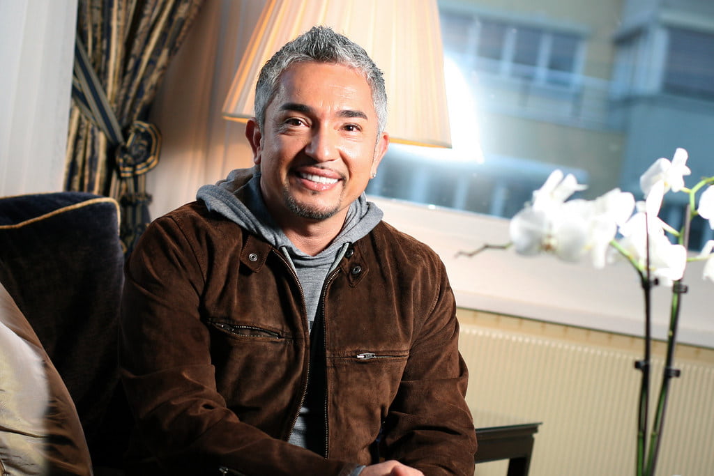 Cesar Millan smiling for the camera with a wall in the background.