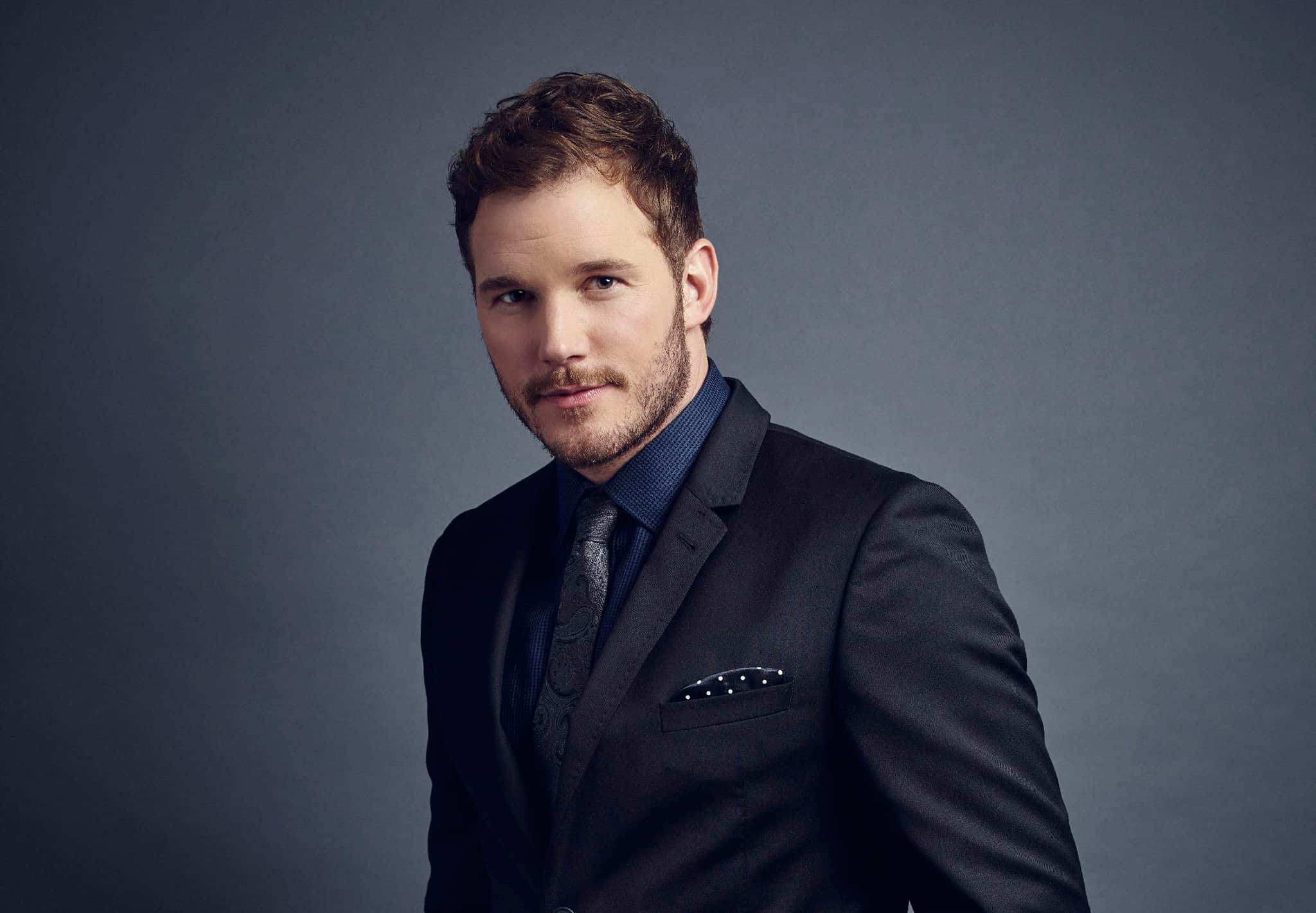 Chris Pratt wearing a black suit, posing in front of a blue background