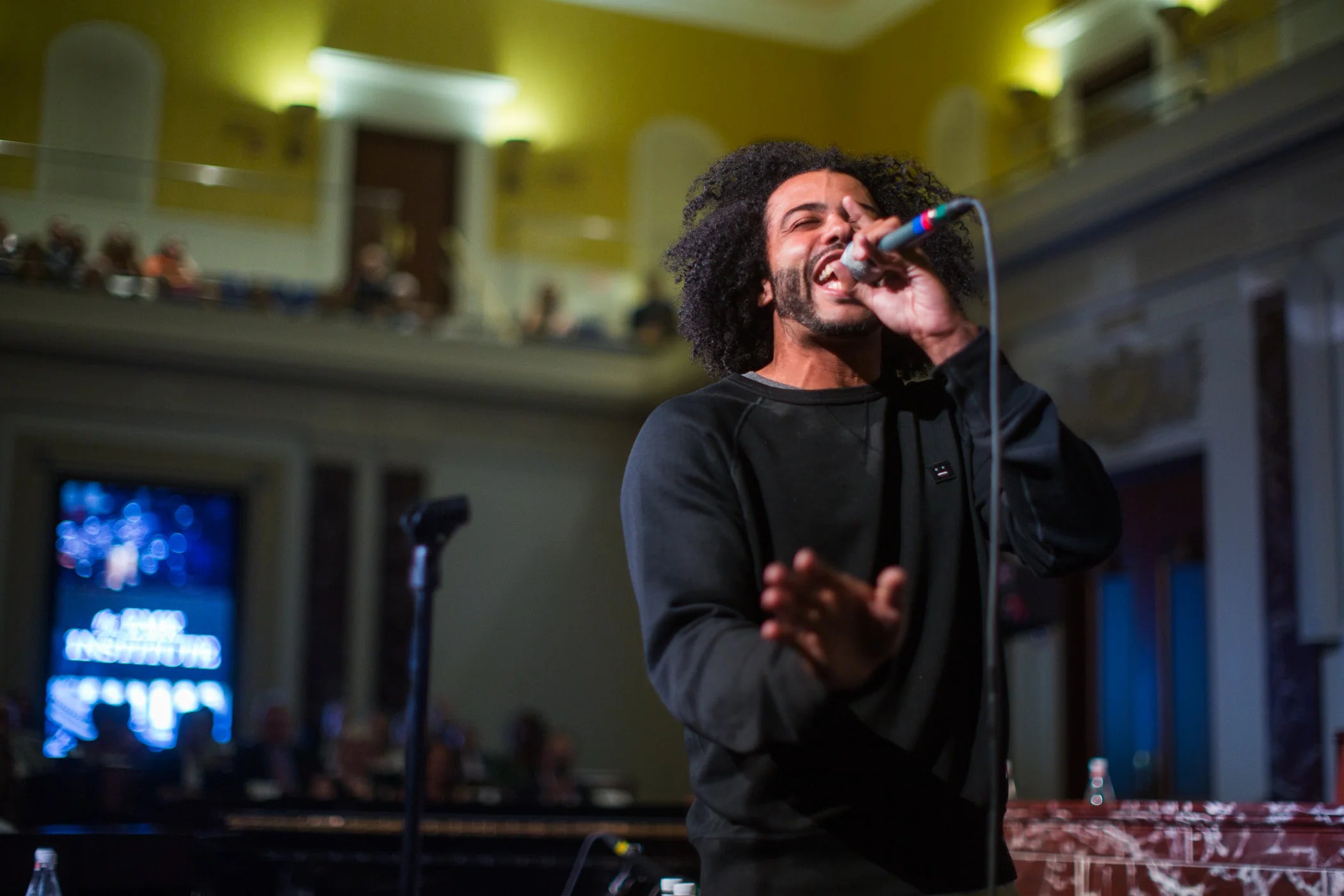 Daveed Diggs on stage at a concert holding a microphone and singing for the audience.