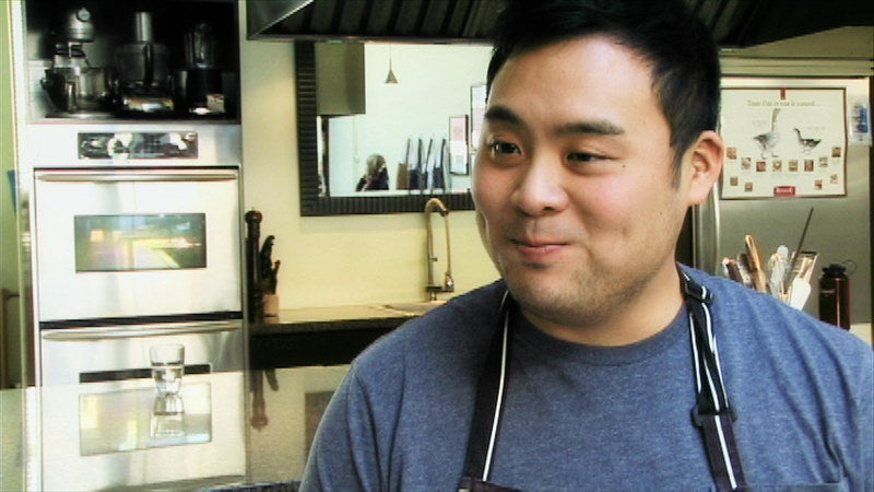 David Chang wearing a blue t-shirt and a black apron, smiling while interacting in his kitchen. 