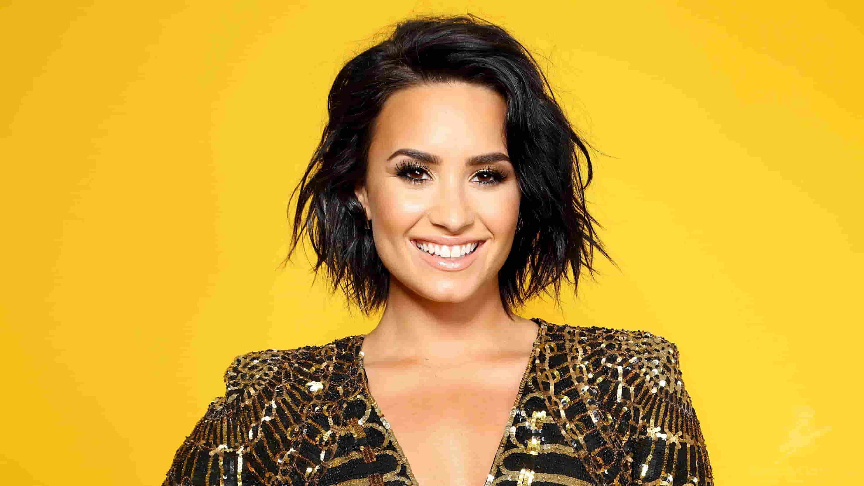 Demi Lovato in a black and gold shimmery dress, smiling while posing in front of a yellow background.