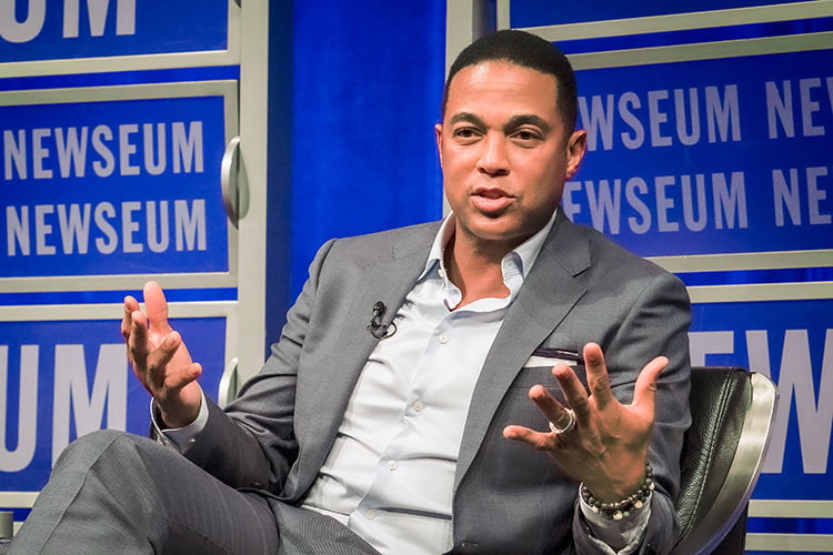 Don Lemon wearing a grey suit, sitting in front of a blue background and addressing the audience at a conference.