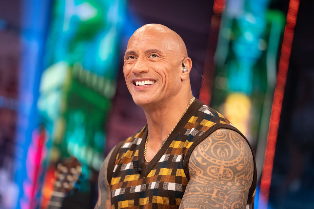 Dwayne in a brown half-sleeve cardigan, sitting in front of a colorful background and smiling