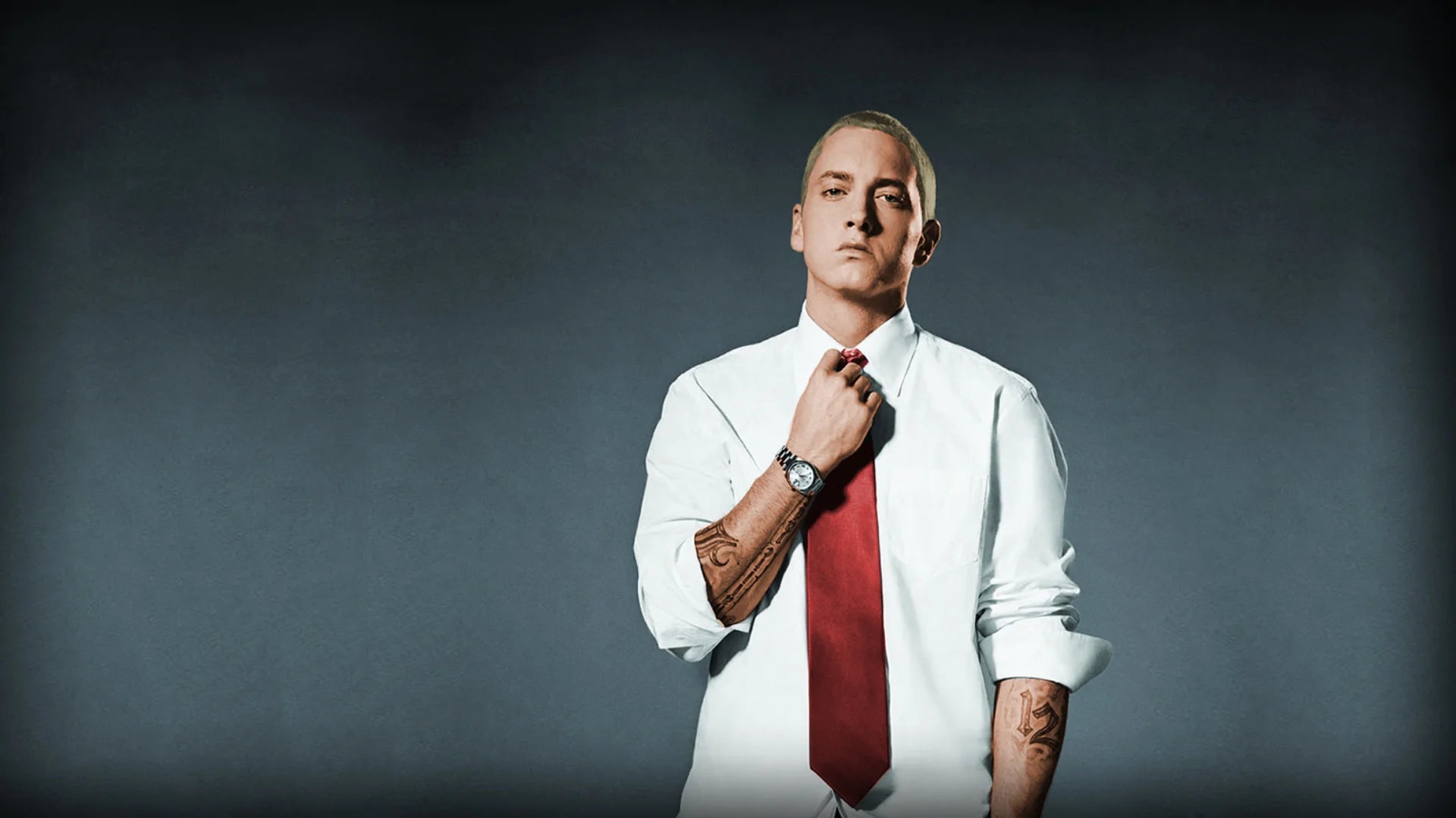 Eminem is dressed in a white shirt and red tie and he is holding the tie with his hand