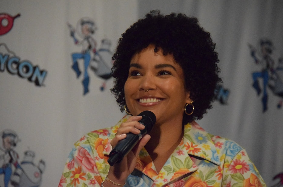 Emmy Raver, wearing a floral t-shirt, holding a mic in her hand and smiling while addressing the audience at a conference