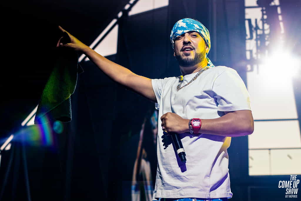 French Montana wearing a hat and holding a microphone with his hand raised.