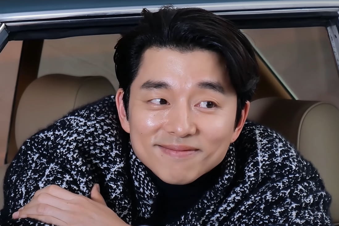 Gong Yoo, wearing a black overcoat and a black high neck, sitting in a car and smiling for the camera.