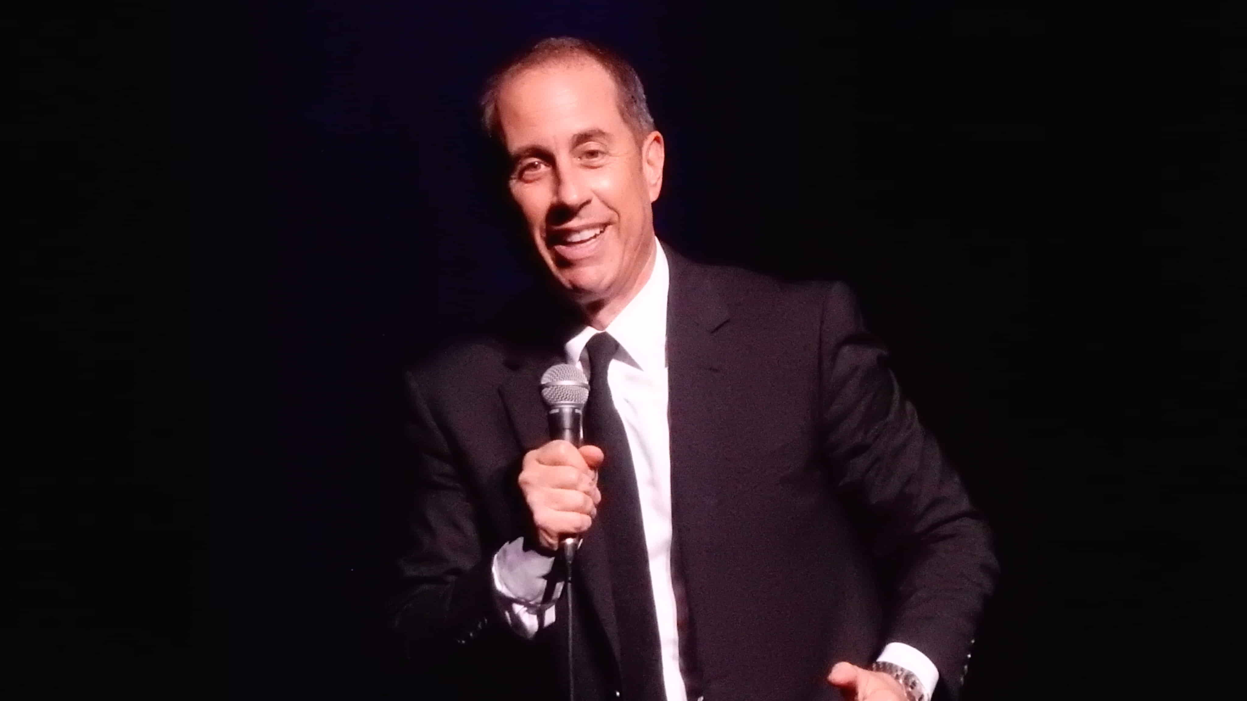 Jerry Seinfeld in a black suit, holding the mic in his hand while smiling and doing his usual stand up routine.