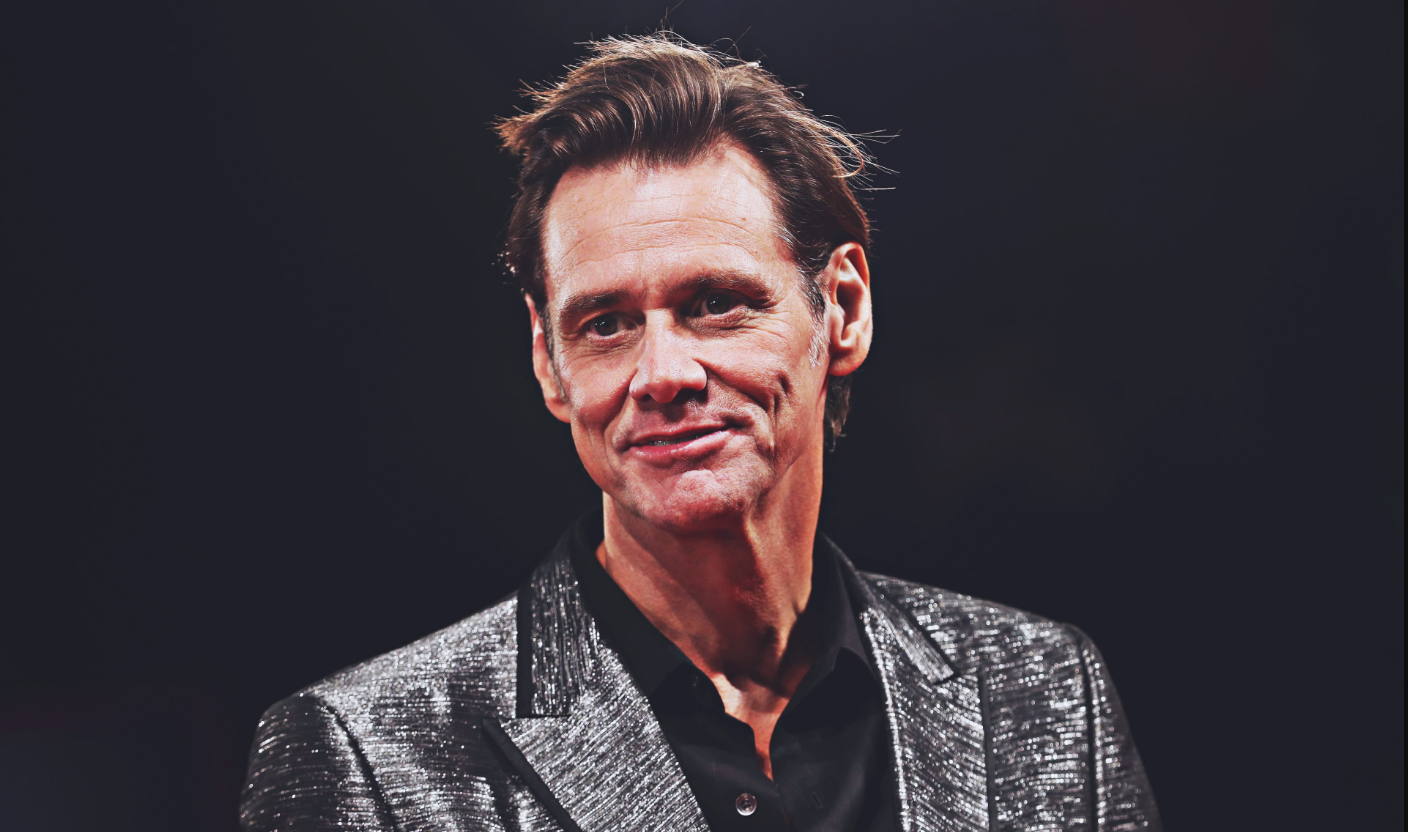 Jim Carrey in a grey suit, standing in front of a black background.