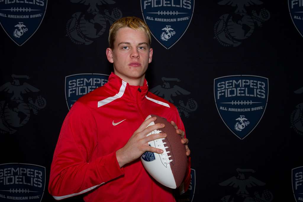 Joe Burrow is in a red Nike zipper jacket, holding rugby football in both hands
