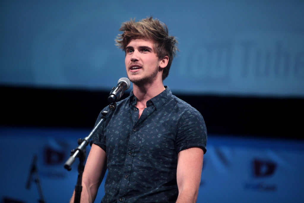 Joey Graceffa in a navy blue shirt addressing the audience through a mic kept in front of him