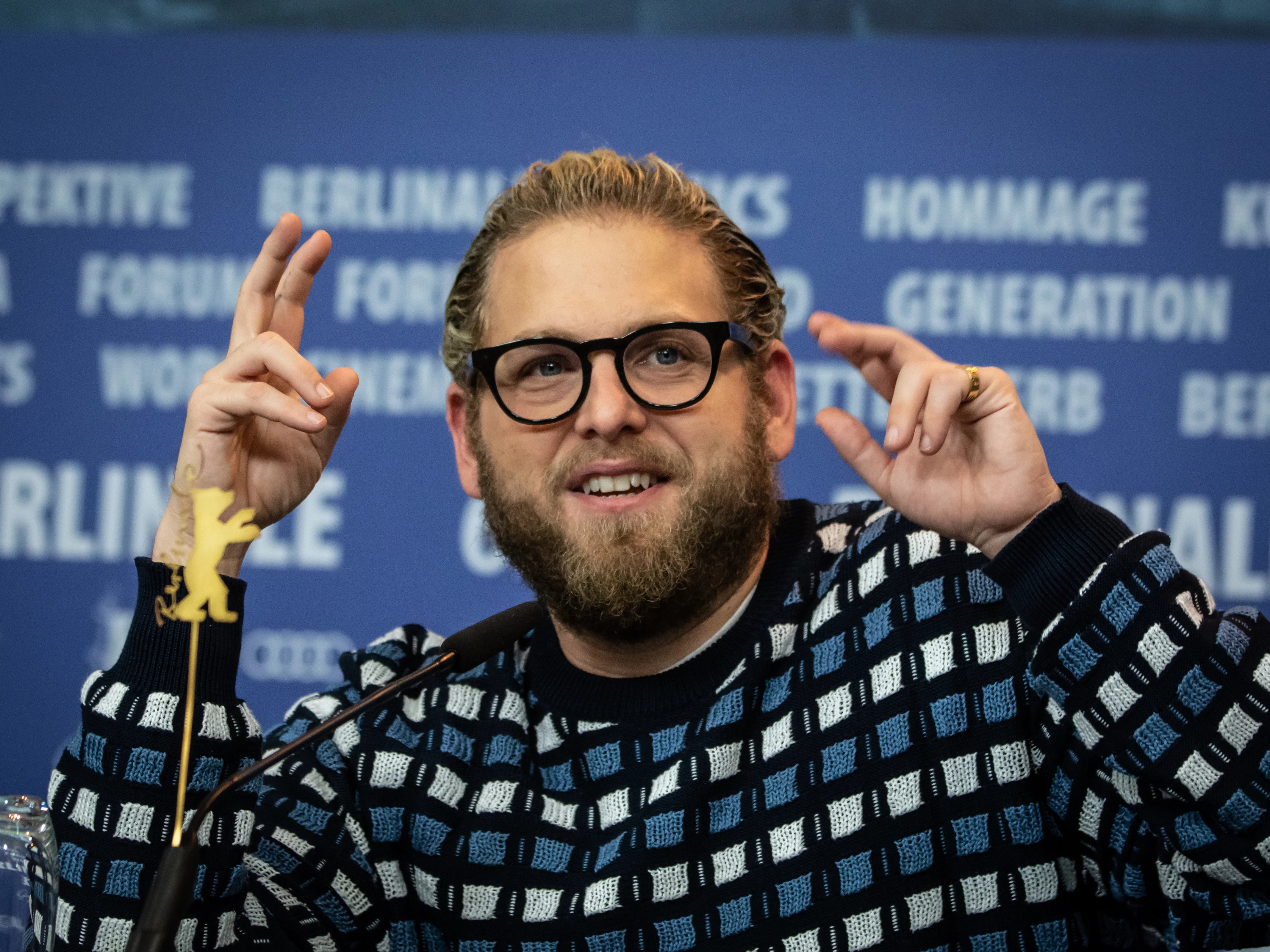 Jonah Hill is at a conference event and he has his hands up in the air with crossed fingers