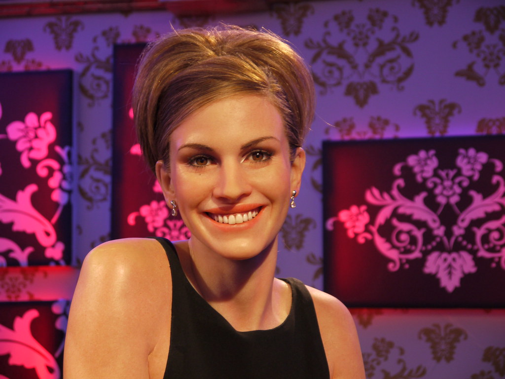 A wax figure of Julia Roberts at Madame Tussauds in London smiles in front of a pink and purple floral background.