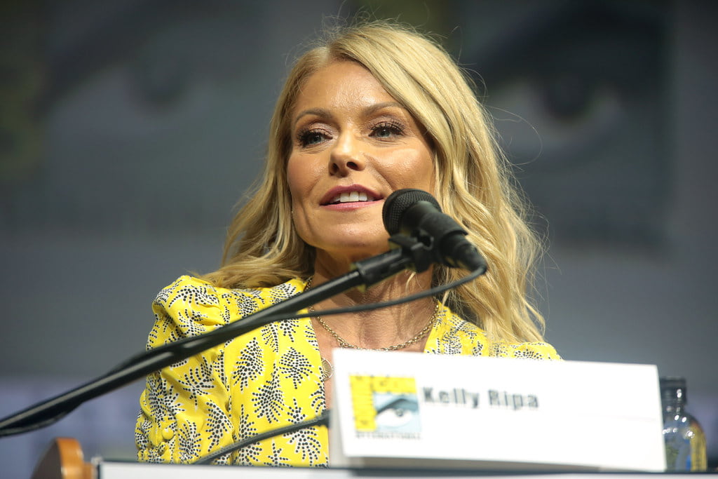 Kelly Ripa in a yellow dress, addressing the audience at the Comic-Con event.