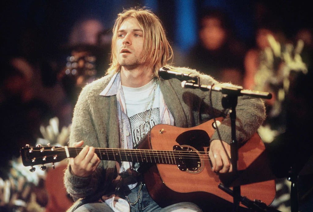 Kurt Cobain in a grey cardigan, playing a guitar and singing at a concert.