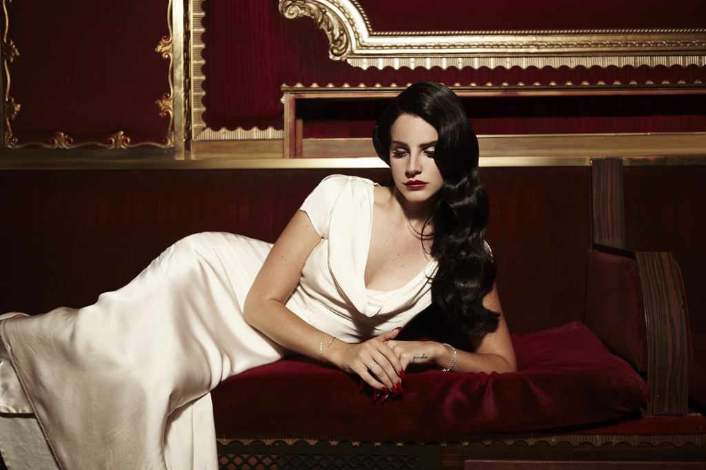 Lana Del Rey in a white dress lying on a red sofa while looking down.