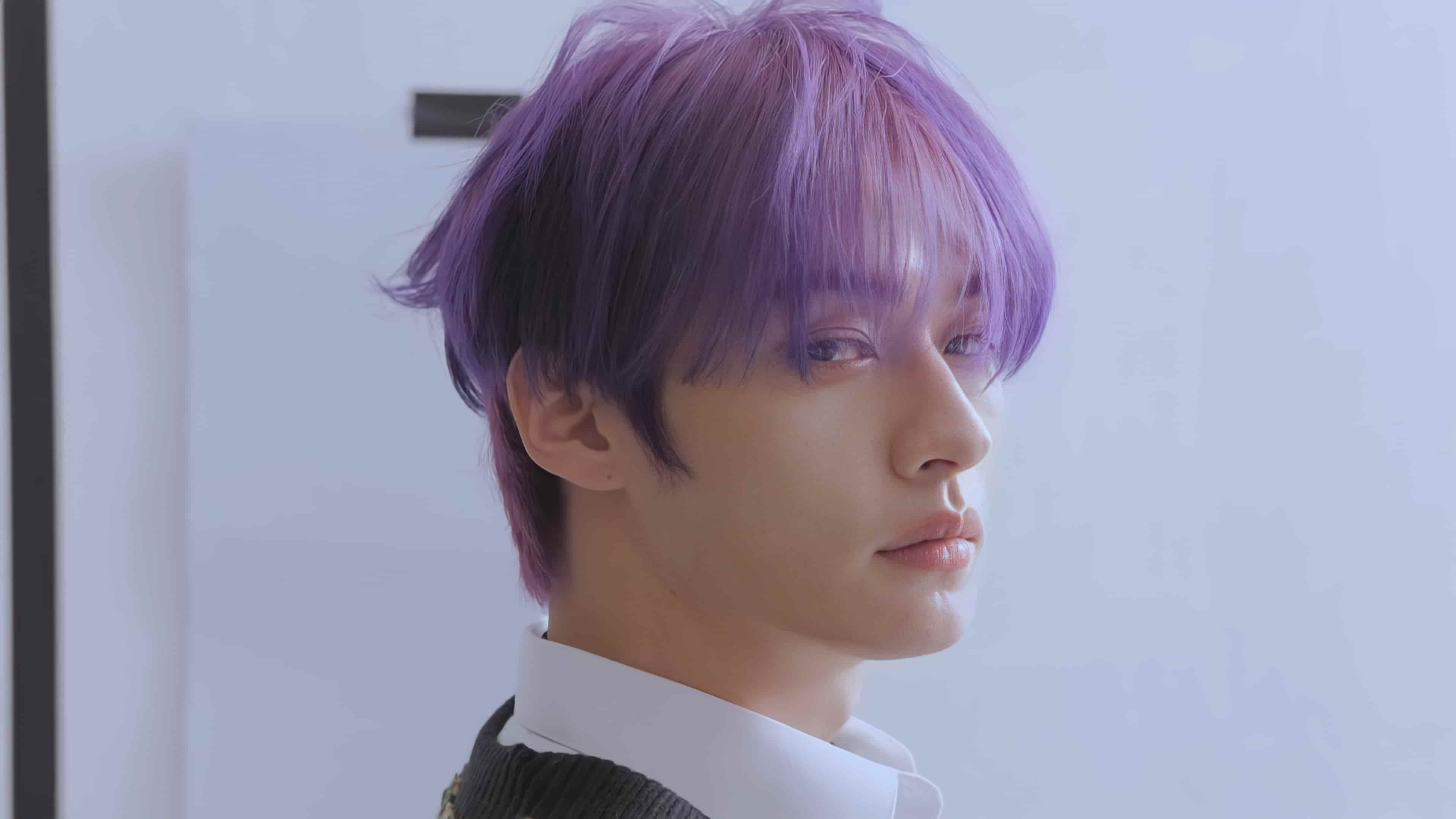 Lee Know with vibrant purple hair, wearing a white shirt and sweater, gazing thoughtfully to the side against a soft, light background.