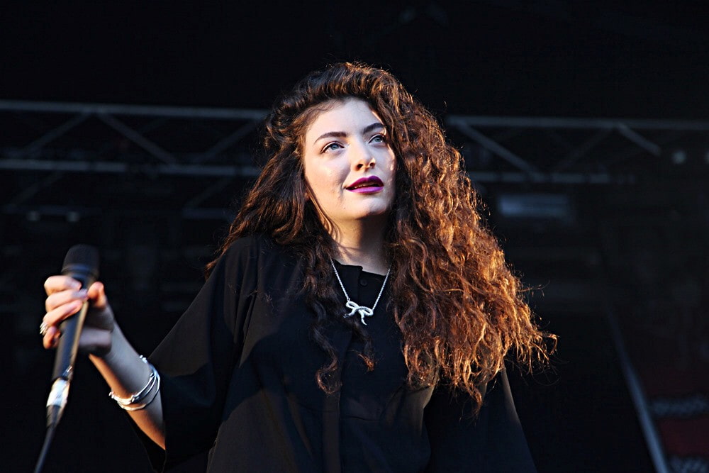 Lorde in a black dress, holding a mic in her right hand and singing at her concert.