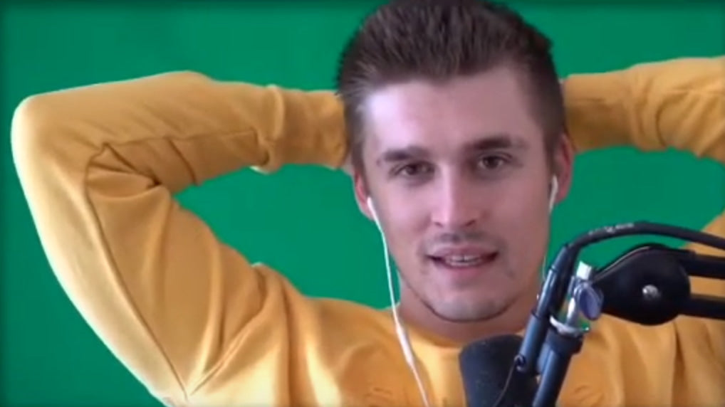 Ludwig Ahgren is sitting in front of a green screen in an orange sweatshirt, with his hands clasped at the back of his head and a microphone kept in front of him