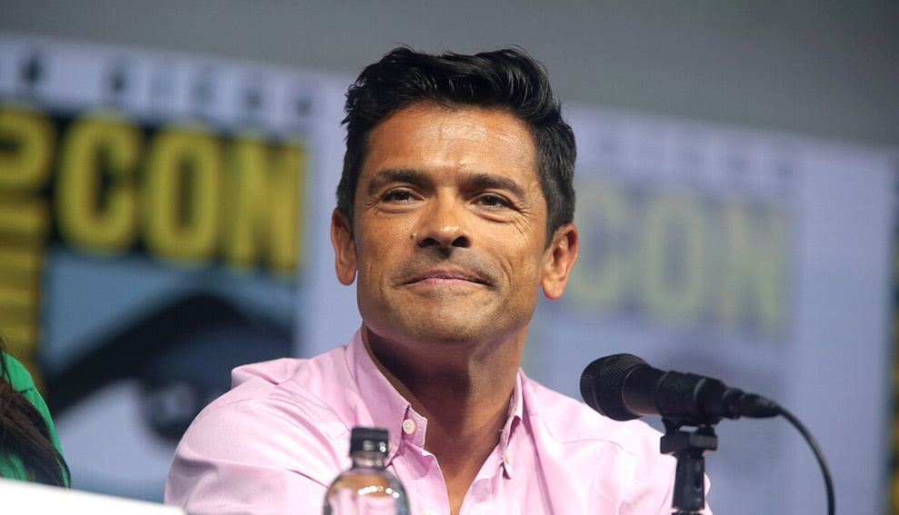 Mark Consuelos in a pink shirt, smiling while addressing the audience at a Comic-Con event.